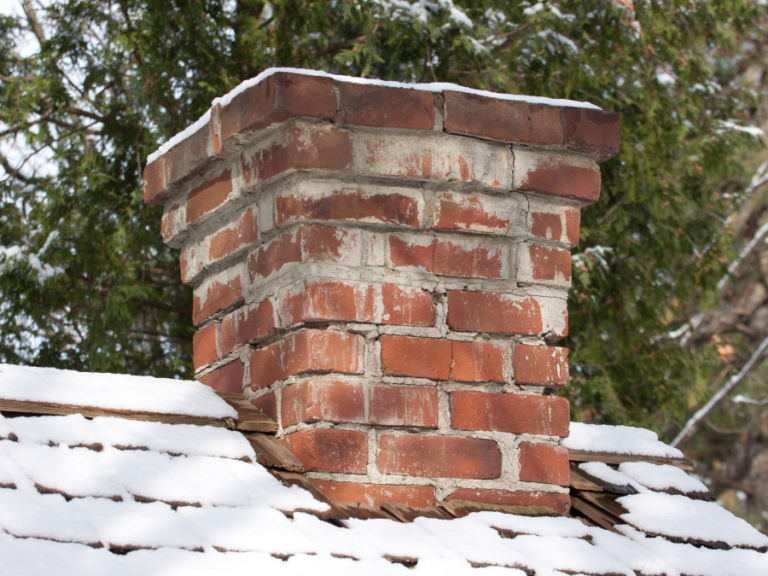 Ceglany komin pokryty śniegiem na tle drzew, co ilustruje potrzebę regularnych przeglądów i e-protokołów kominiarskich.